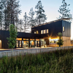A beautifully designed modern home with a striking facade, featuring large windows and a combination of wood and metal elements. The house is set in a natural landscape, surrounded by trees and greenery.