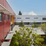 Exterior view of Wing Luke Elementary School, featuring a modern architectural design with colorful panels and large windows. The building is surrounded by landscaped areas, including trees and play spaces. The entrance is welcoming, with a covered walkway and signage that highlights the school's name, reflecting a vibrant and engaging environment for students.