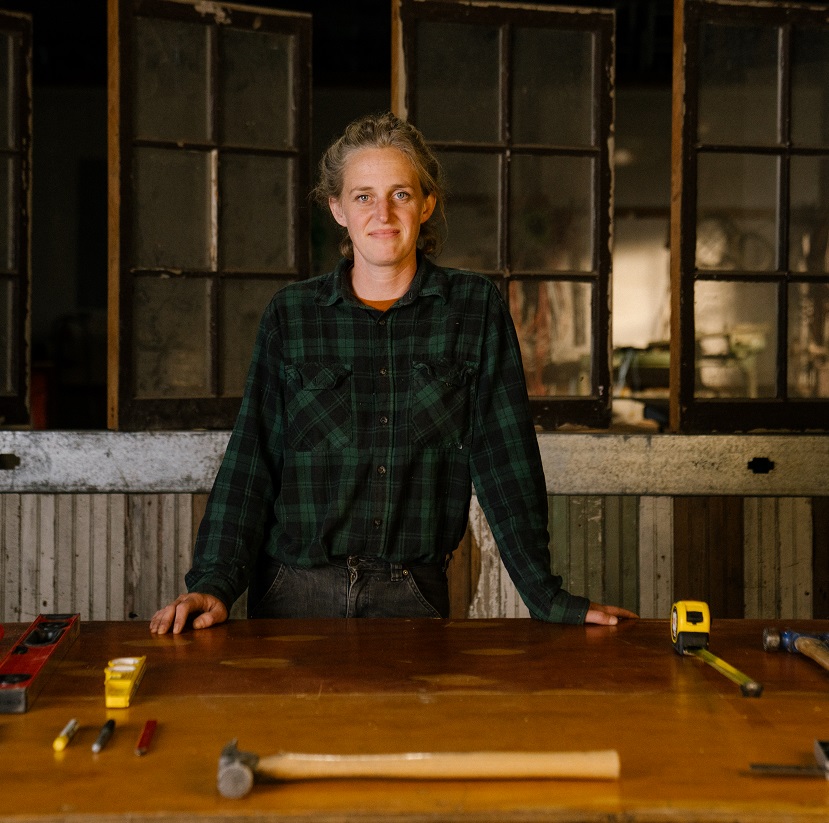 Color headshot of a feminine person in a workshop, hands perched on a work bench in front of a many-paneled window.