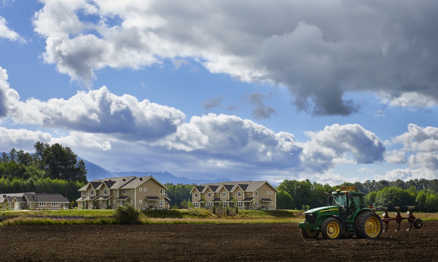 view of farmland with large tractor tilling a field, with blue sky, clouds, and beige housing in background