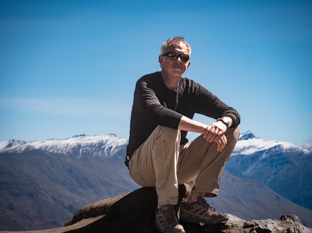 Crouching man on mountain peak in khaki pants, dark longsleeve short, arms perched on knees, wearing sunglasses. The background is a series of snowcapped mountain peaks