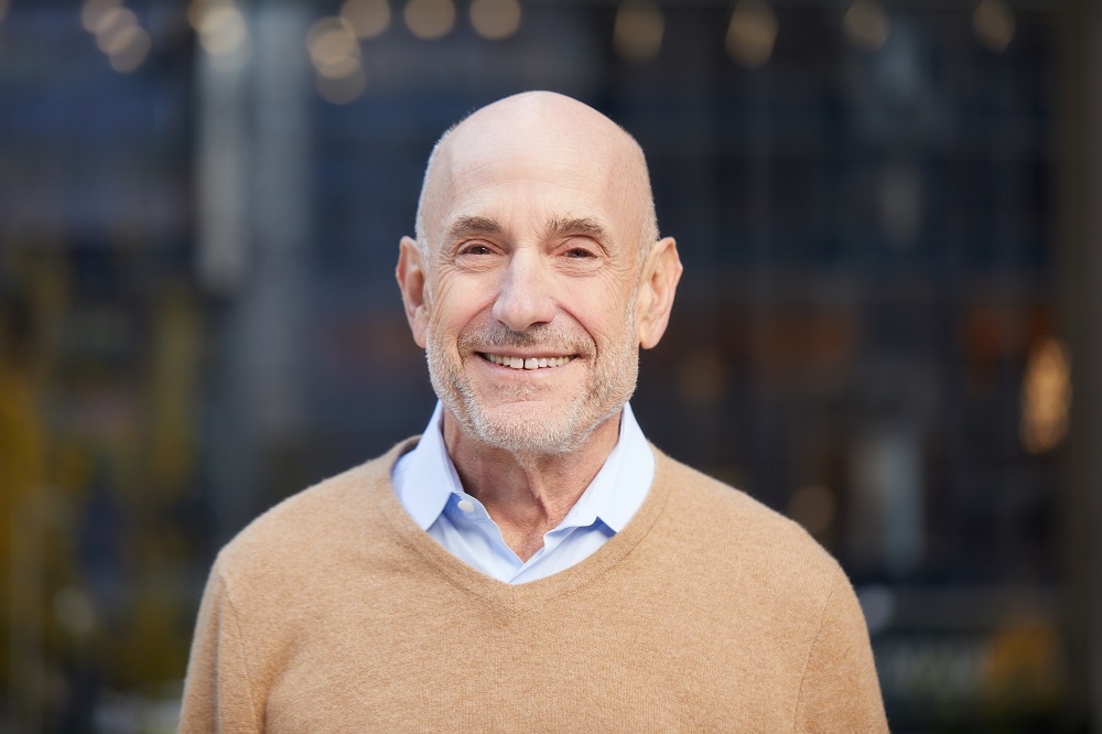 Bald white man in a beige sweater with light blue collar peeking out, smiling and standing in front of an out-of-focus outdoor background